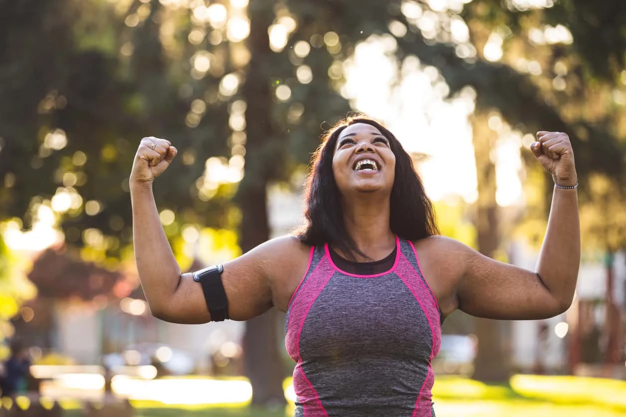 A woman in athletic wear smiles joyfully with her arms raised in victory while standing outdoors in a sunlit park, celebrating a fitness achievement.