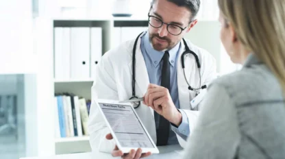 A doctor wearing a white coat and stethoscope shows and explains a document on a tablet to a patient in an office setting.