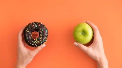 Two hands against an orange background: the left hand holds a chocolate donut with colorful sprinkles, and the right hand holds a green apple.