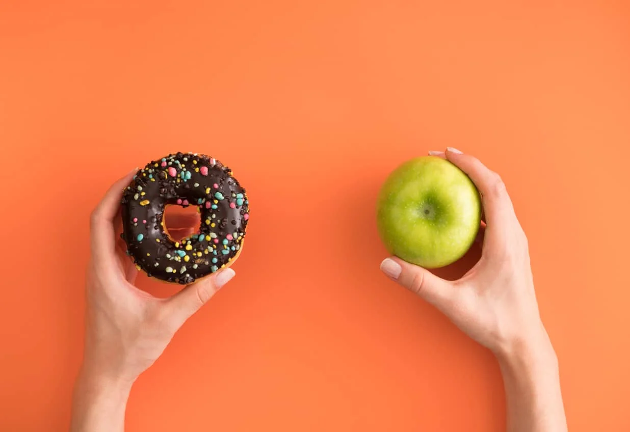 Two hands against an orange background: the left hand holds a chocolate donut with colorful sprinkles, and the right hand holds a green apple.