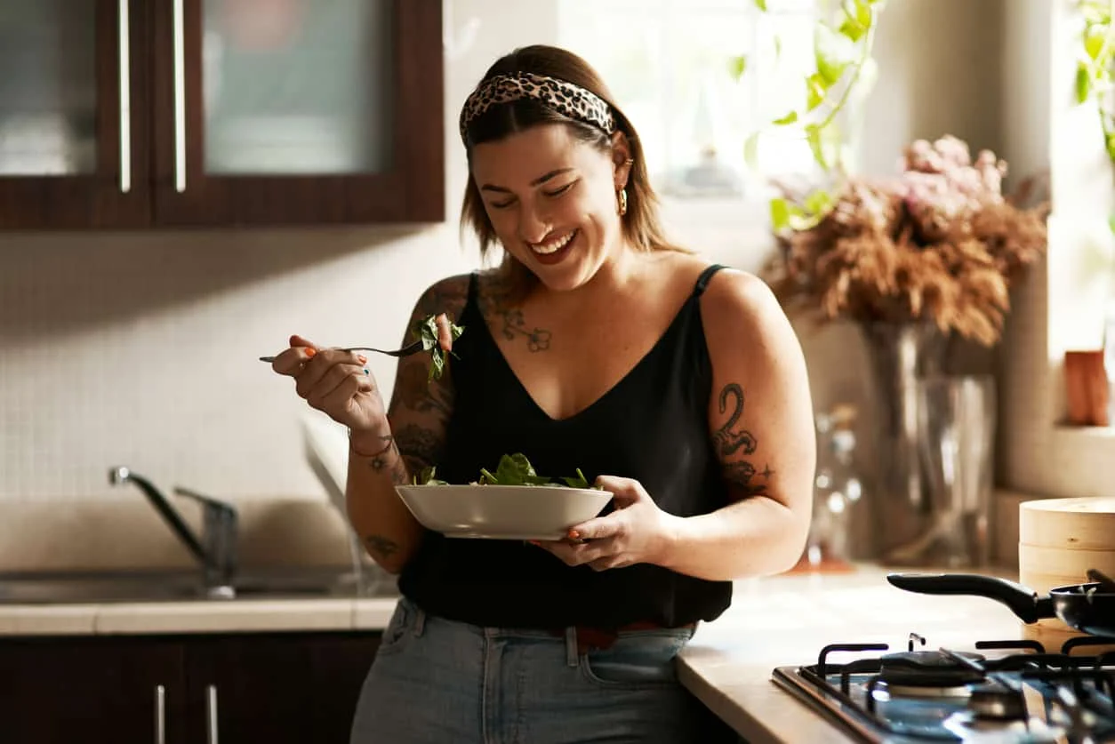 A smiling woman with tattoos stands in a kitchen, holding a bowl of salad and a fork. She is wearing a black tank top, jeans, and a leopard-print headband. Sunlight streams in through the window behind her.