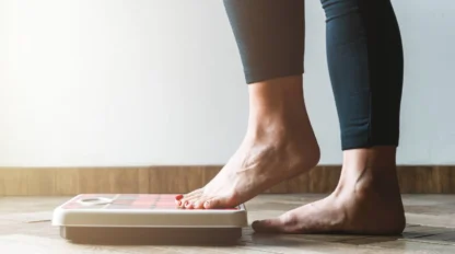 A person wearing dark leggings is standing barefoot on a digital bathroom scale placed on a wooden floor, checking their weight.