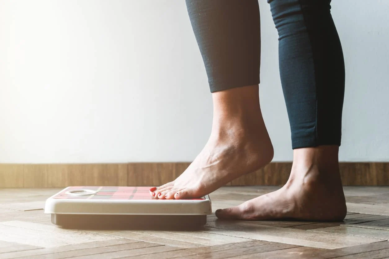 A person wearing dark leggings is standing barefoot on a digital bathroom scale placed on a wooden floor, checking their weight.