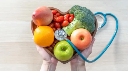 Hands holding a heart-shaped bowl filled with fruits and vegetables, including an apple, orange, tomato, peach, and broccoli, with a stethoscope placed on top, symbolizing healthy eating and heart health.