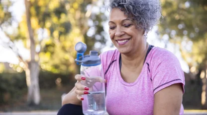 A smiling woman with short curly gray hair sits outdoors, holding a clear water bottle. She wears a pink athletic shirt and has red painted nails. Trees and greenery are blurred in the background.