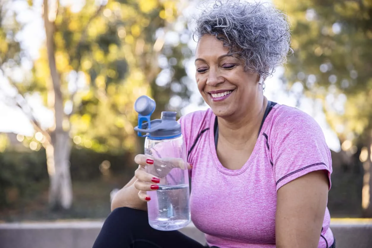 A smiling woman with short curly gray hair sits outdoors, holding a clear water bottle. She wears a pink athletic shirt and has red painted nails. Trees and greenery are blurred in the background.