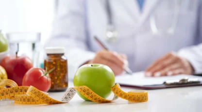 A green apple and measuring tape are on a table, with a blurred figure in a white coat writing on a clipboard in the background. Nearby are medications, a glass of water, and other fruits.