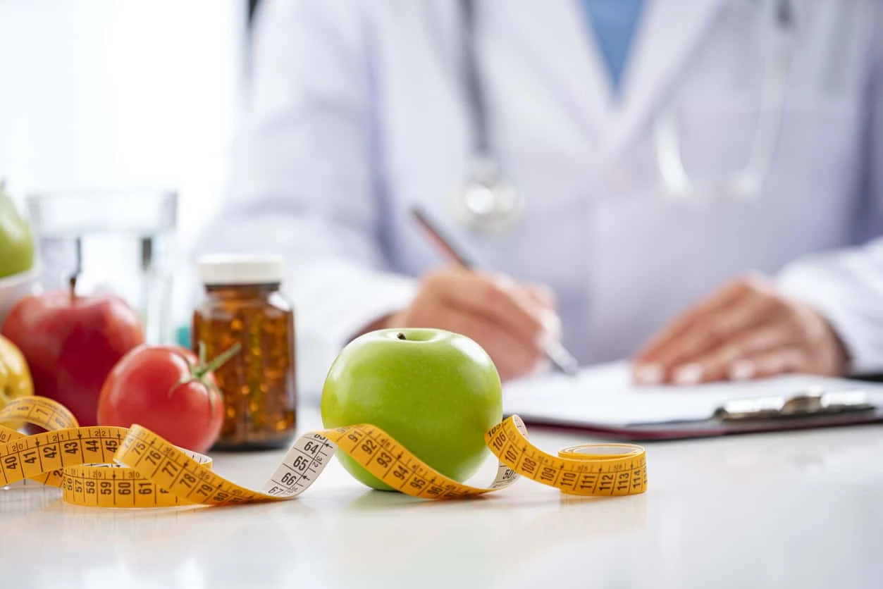 A green apple and measuring tape are on a table, with a blurred figure in a white coat writing on a clipboard in the background. Nearby are medications, a glass of water, and other fruits.