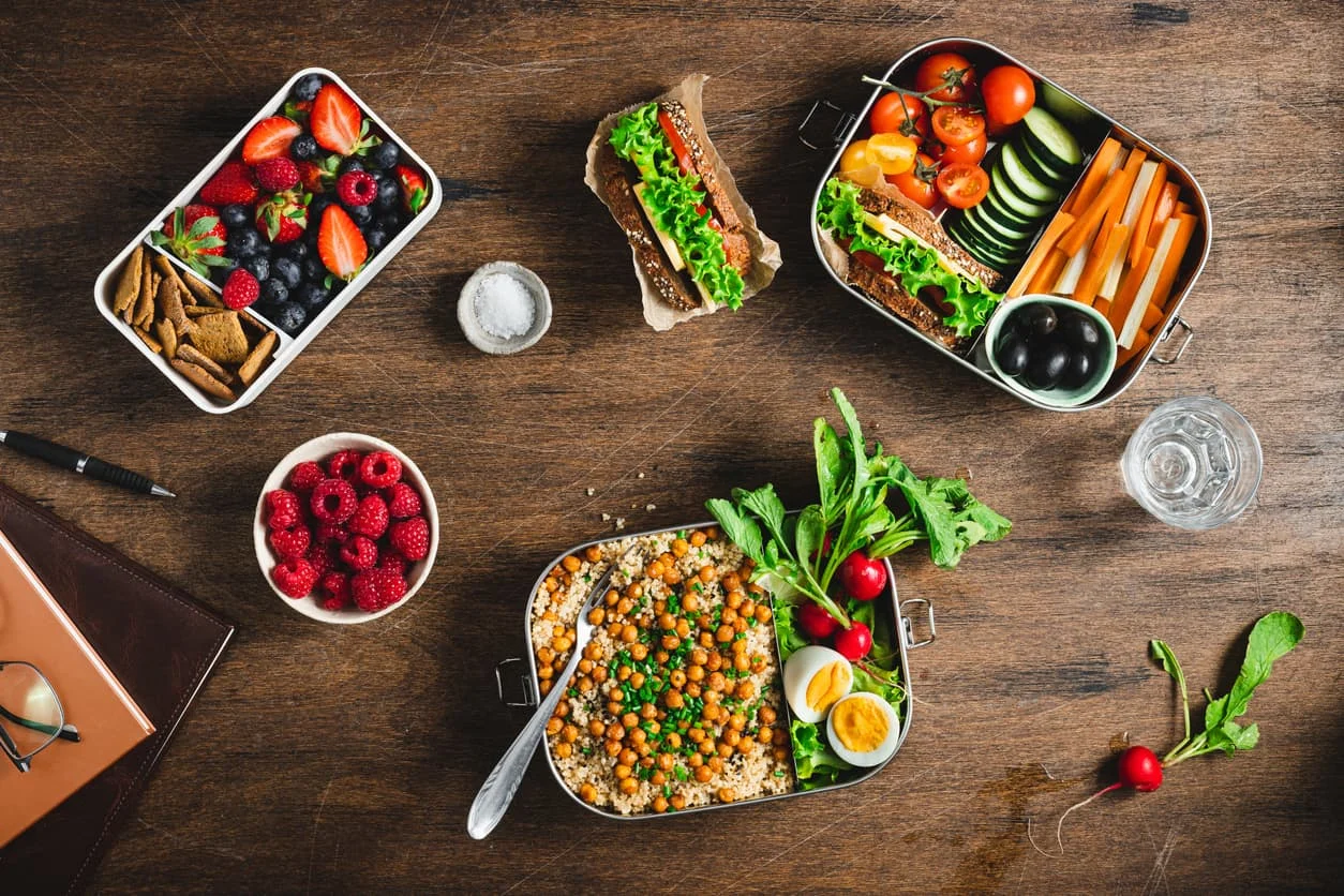 A wooden table with containers of fresh food, including salad with chickpeas and eggs, sliced vegetables, a sandwich, berries, crackers, and a glass of water. Radishes and greens are scattered on the table.