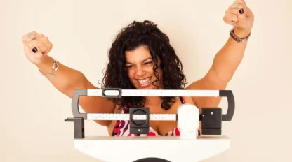 A woman with curly hair smiles and raises her fists in excitement while standing on a mechanical scale, celebrating her progress.