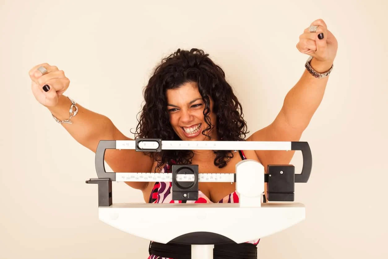 A woman with curly hair smiles and raises her fists in excitement while standing on a mechanical scale, celebrating her progress.