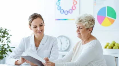 A healthcare professional in a white coat shows a clipboard to an older woman in a bright office, with colorful charts on the wall in the background.