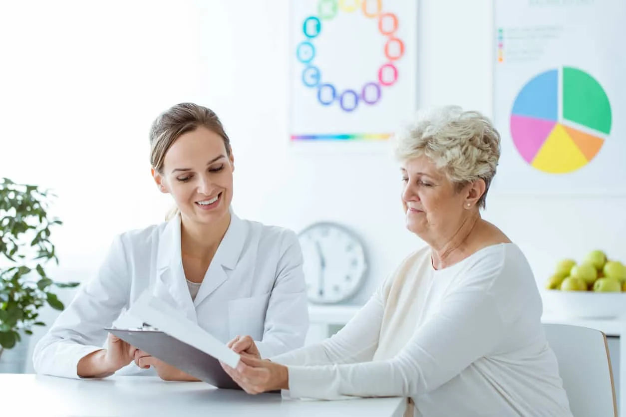 A healthcare professional in a white coat shows a clipboard to an older woman in a bright office, with colorful charts on the wall in the background.