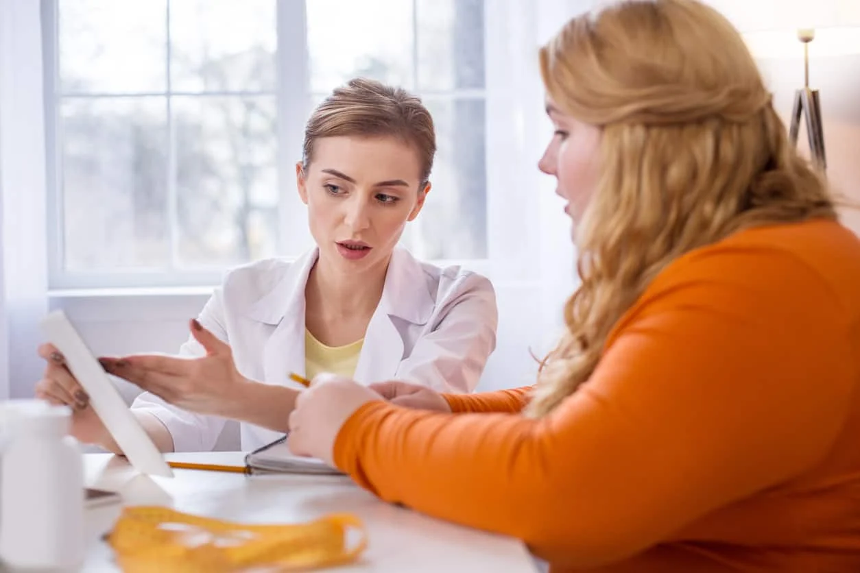 A doctor discusses information on a tablet with a patient at a desk in a bright office.