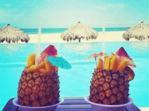 Two hollowed pineapples filled with drinks and garnished with fruit and umbrellas sit by a blue pool, with straw beach huts and sand in the background.
