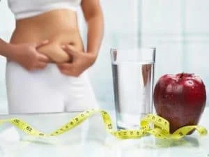 A person pinches their abdominal skin; in the foreground are a glass of water, a red apple, and a yellow measuring tape on a glass surface.