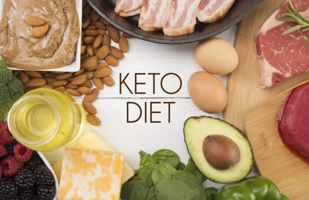 Assorted keto-friendly foods including meat, eggs, avocado, cheese, spinach, almonds, nut butter, oil, and berries, arranged around the words "Keto Diet" on a white surface.