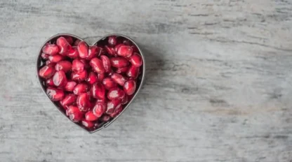 A heart-shaped container filled with pomegranate seeds sits on a light gray wooden surface.