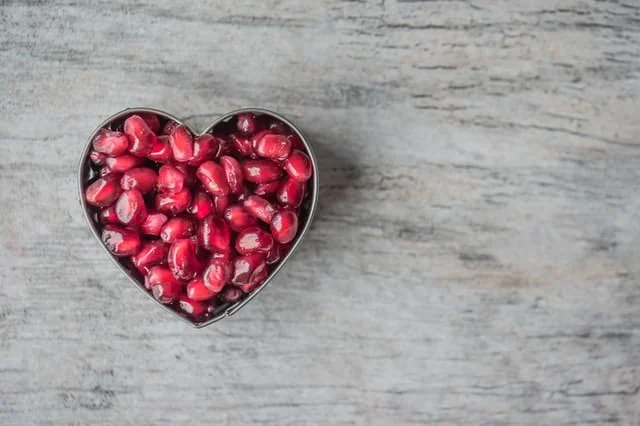A heart-shaped container filled with pomegranate seeds sits on a light gray wooden surface.