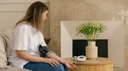A woman sits on a couch at home, measuring her blood pressure with a digital monitor on her arm. A vase of flowers and medical items are on a wooden table in front of her, next to a stone fireplace.
