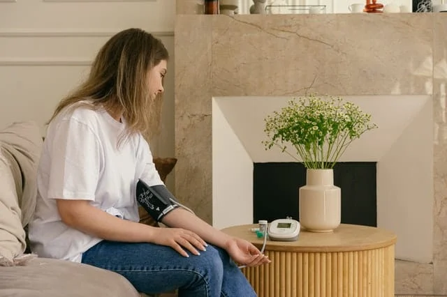 A woman sits on a couch at home, measuring her blood pressure with a digital monitor on her arm. A vase of flowers and medical items are on a wooden table in front of her, next to a stone fireplace.