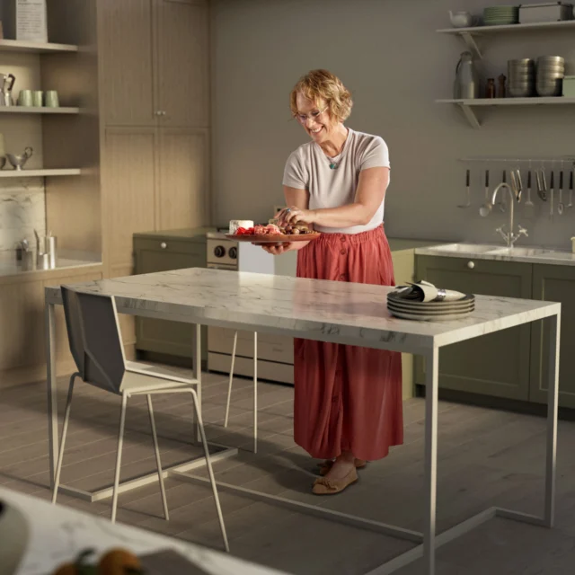 A woman stands in a kitchen, cutting vegetables on a counter. The kitchen has green cabinets, open shelving with plates, and a white countertop.