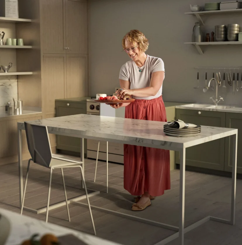 A woman stands in a kitchen, cutting vegetables on a counter. The kitchen has green cabinets, open shelving with plates, and a white countertop.