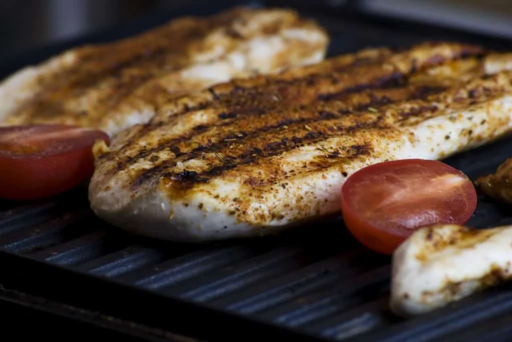 Seasoned chicken breasts and sliced tomatoes are cooking on a ridged grill pan, with visible grill marks on the chicken.