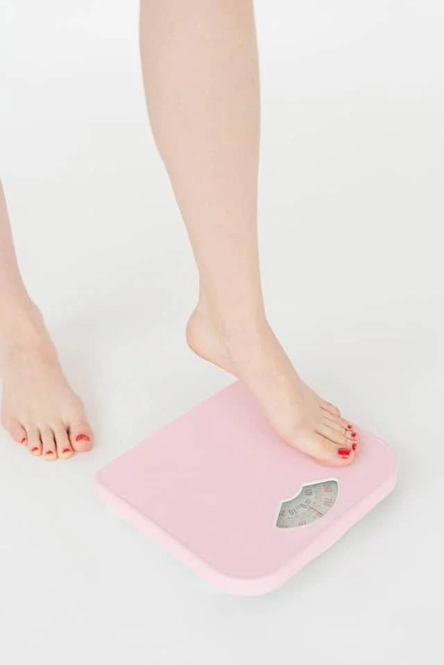 A person with painted toenails steps onto a pink bathroom scale on a white floor, preparing to check their weight.