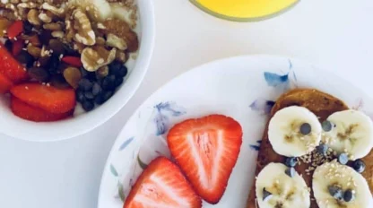 A bowl of yogurt with fruit and nuts, a glass of orange juice, and a plate with toast topped with banana slices, chocolate chips, and two strawberry halves.