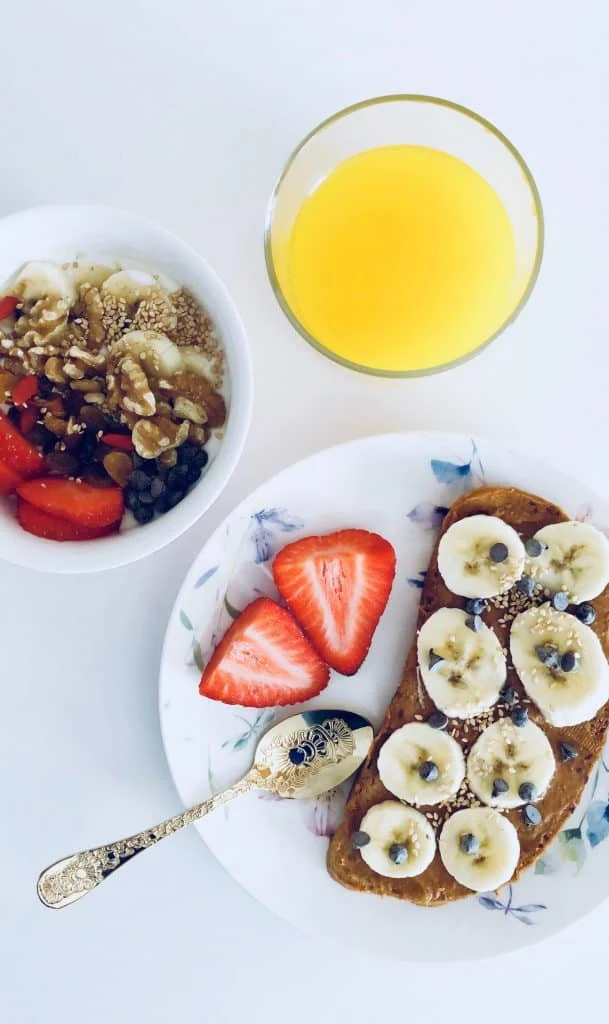 A bowl of yogurt with fruit and nuts, a glass of orange juice, and a plate with toast topped with banana slices, chocolate chips, and two strawberry halves.