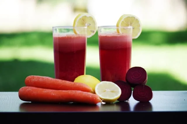 Two glasses of red juice with lemon slices on the rim, placed on a table with fresh carrots, a whole lemon, a halved lemon, and sliced beets in front. The background is green and blurred.