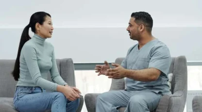 A woman in casual clothes sits on a chair talking to a man in medical scrubs. They are facing each other in a bright, modern room, engaged in conversation.