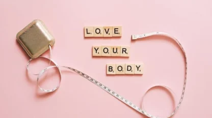 A measuring tape and square container on a pink background, with Scrabble tiles spelling “LOVE YOUR BODY” arranged in the center.