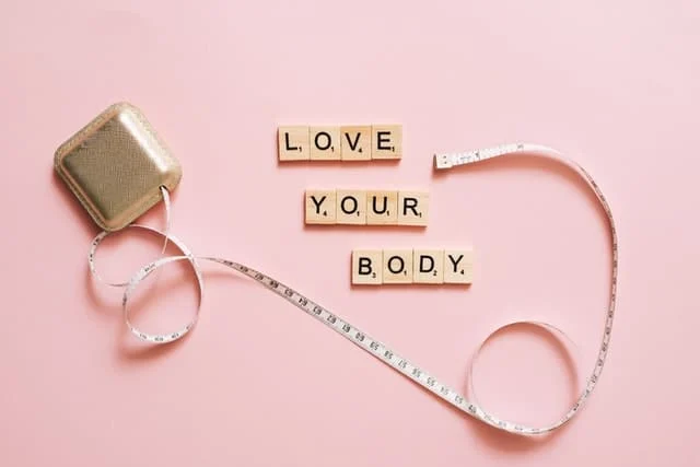 A measuring tape and square container on a pink background, with Scrabble tiles spelling “LOVE YOUR BODY” arranged in the center.
