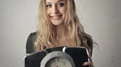 A young woman with long, light brown hair is smiling and holding a black analog bathroom scale in front of her against a plain background.