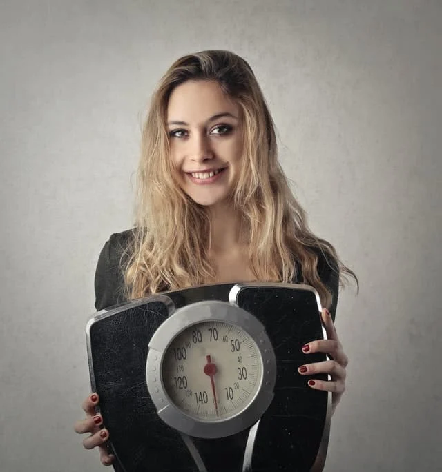 A young woman with long, light brown hair is smiling and holding a black analog bathroom scale in front of her against a plain background.