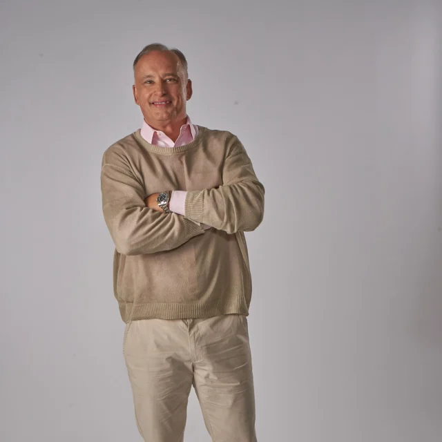 Man in a tan sweater and light pants standing with arms crossed against a plain gray background.