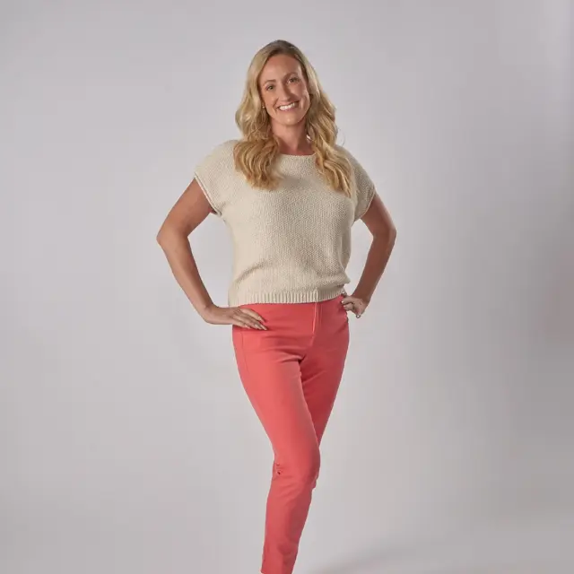 A woman with long blonde hair, wearing a short-sleeve cream sweater, coral pants, and sandals, stands smiling against a plain white background.