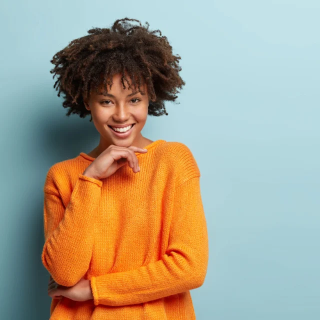 Person with curly hair wearing an orange sweater smiles against a blue background.