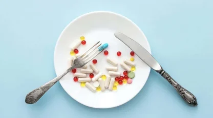 A white plate with capsules and pills arranged on it, with a fork and knife placed on either side, against a pale blue background.
