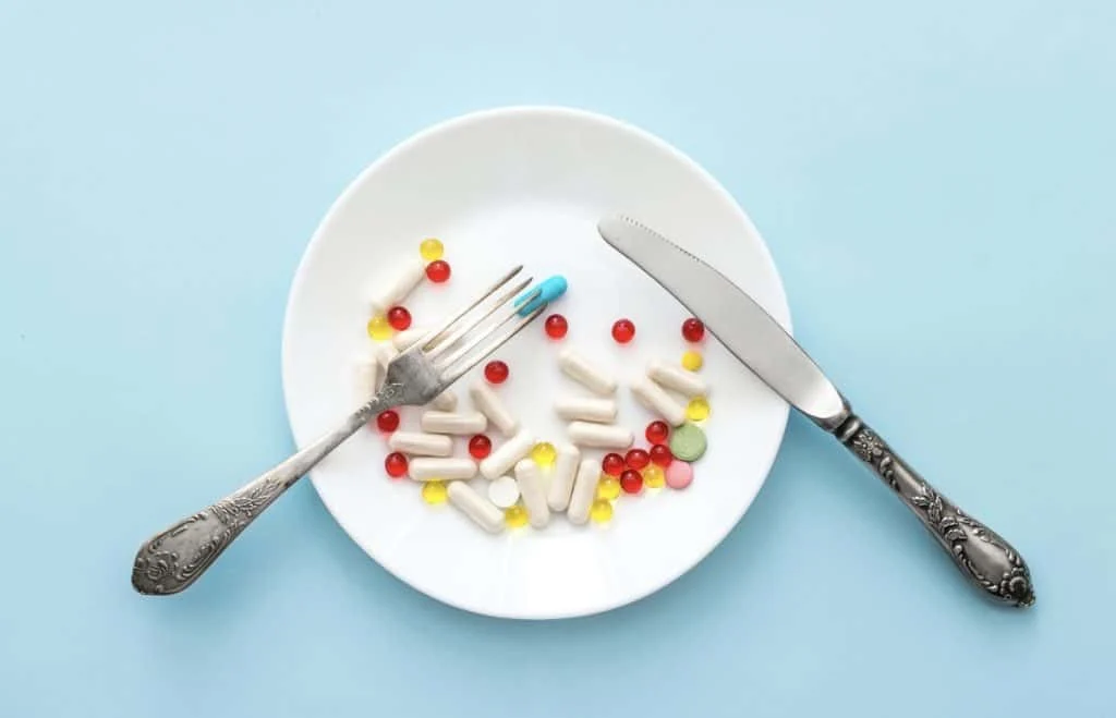 A white plate with capsules and pills arranged on it, with a fork and knife placed on either side, against a pale blue background.
