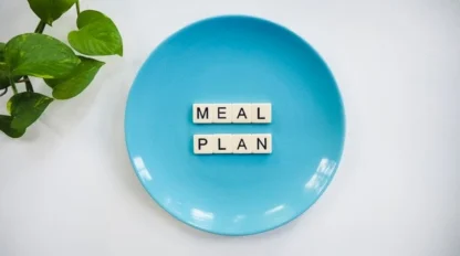 A blue plate with letter tiles spelling "MEAL PLAN" on it, placed on a white surface next to a green leafy plant.