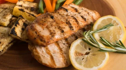 Grilled chicken breast with grill marks, served with grilled vegetables, carrot sticks, green beans, and garnished with lemon slices and rosemary on a wooden plate.