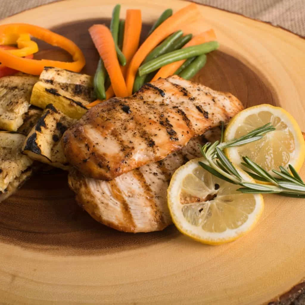 Grilled chicken breast with grill marks, served with grilled vegetables, carrot sticks, green beans, and garnished with lemon slices and rosemary on a wooden plate.