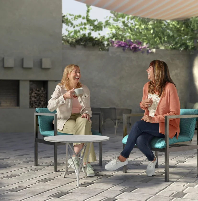 Two women sit and chat on teal armchairs in a sunlit patio. A small table is between them, and there's a fireplace in the background.