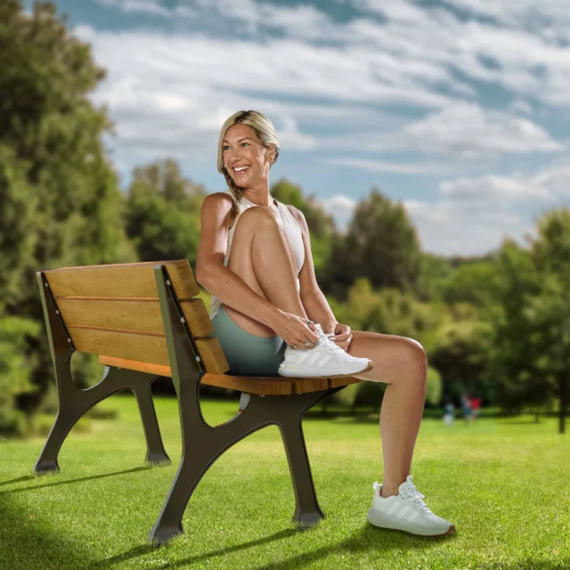 A person sitting on a park bench with legs crossed, smiling, surrounded by green grass and trees under a partly cloudy sky.