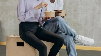Two people sit together on a wooden box, eating from bowls and smiling in a casual indoor setting with a gray concrete wall behind them.