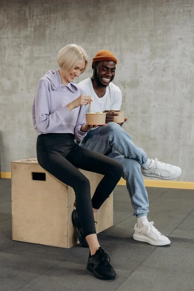 Two people sit together on a wooden box, eating from bowls and smiling in a casual indoor setting with a gray concrete wall behind them.