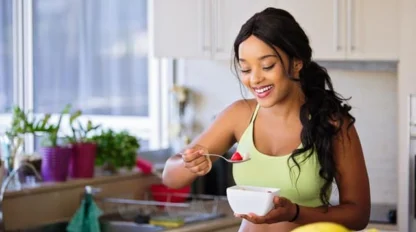 A woman in a green sports bra smiles while eating fruit from a white bowl in a bright kitchen with potted plants and bananas visible on the counter.